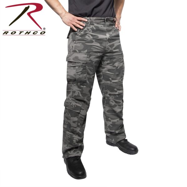 Cargo pants with a camouflage pattern are being worn by a person standing with hands on hips in a neutral background emphasizing the pants and black boots. Rothco logo is visible.