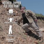A person in a tan jacket and pants crouches on rocky terrain reaching for the ground surrounded by grass and rocky soil showcasing outdoor activity and functionality of clothing