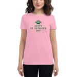 A woman stands wearing a pink t-shirt featuring green text and a graphic that reads Happy St. Patrick's Day  celebrating the holiday in a casual  festive context.