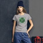 A young woman wearing a gray t-shirt with a green shamrock design stands against a textured dark wall holding her arm. She has long hair and a dark beanie on her head.
