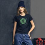 A young woman stands against a textured wall wearing a black t-shirt with a green shamrock design celebrating St. Patrick's Day while wearing a dark beanie with casual denim pants