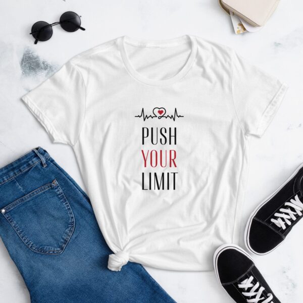 A white t-shirt displays the text PUSH YOUR LIMIT alongside a heartbeat graphic. It is styled with knotted detailing and placed near blue jeans and black sneakers on a light surface.