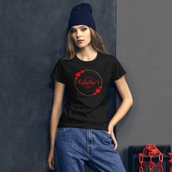 A woman stands against a textured dark wall wearing a black t-shirt with a red and gold design that says Happy Valentine’s Day alongside red heart graphics while wearing a blue beanie.