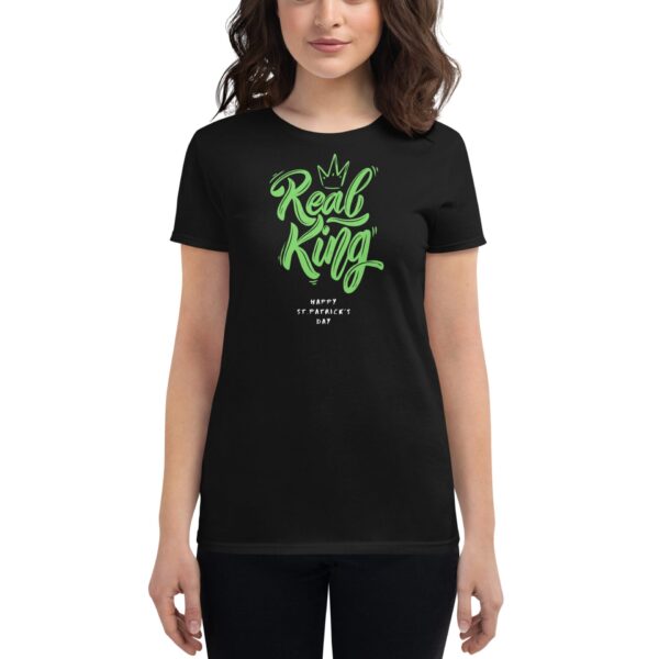 A woman is wearing a black t-shirt featuring the text "Real King" in green cursive with a crown design above it and the phrase "Happy St. Patrick's Day" below.