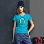 A young woman stands confidently wearing a bright turquoise t-shirt with a rainbow design and text that says Happy St. Patrick's Day against a dark textured background.