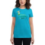 A woman models a bright turquoise t-shirt featuring the text "HAPPY ST. PATRICK'S DAY" with a cartoon character holding a cake and surrounded by decorative elements.