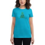 A young woman is wearing a bright turquoise t-shirt featuring text that reads GOOD VIBES and HAPPY ST. PATRICK'S DAY with a rainbow graphic  standing against a neutral background.