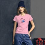 A young woman wearing a pink T-shirt with a heart design stands against a dark wall holding her hand on her hip while wearing a blue beanie.