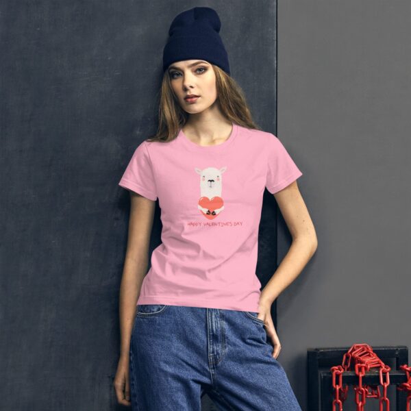 A woman stands against a dark gray wall wearing a pink t-shirt featuring a cartoon llama holding a heart with the text Happy Valentine's Day while wearing a blue beanie and casual jeans.