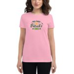 A woman wears a pink t-shirt featuring the text "my first St. Patrick's Day" in green letters with decorative elements  standing against a neutral background.