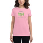A woman poses wearing a pink T-shirt featuring green text that reads limited edition 1974 classic happy st.patrick's day against a neutral background.
