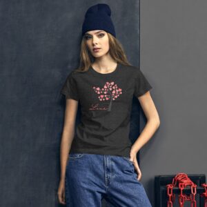 A young woman stands confidently wearing a dark t-shirt with a pink heart tree design and the word "Love" beneath it in a casual setting with a gray wall and chains nearby.
