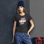 A young woman stands confidently against a dark wall wearing a black t-shirt featuring the word "LOVE" with floral designs and blue jeans while sporting a navy beanie.