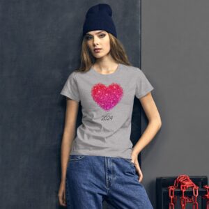 A young woman stands against a gray wall wearing a gray t-shirt featuring a sparkling heart design and the year 2024 with denim jeans and a navy beanie.