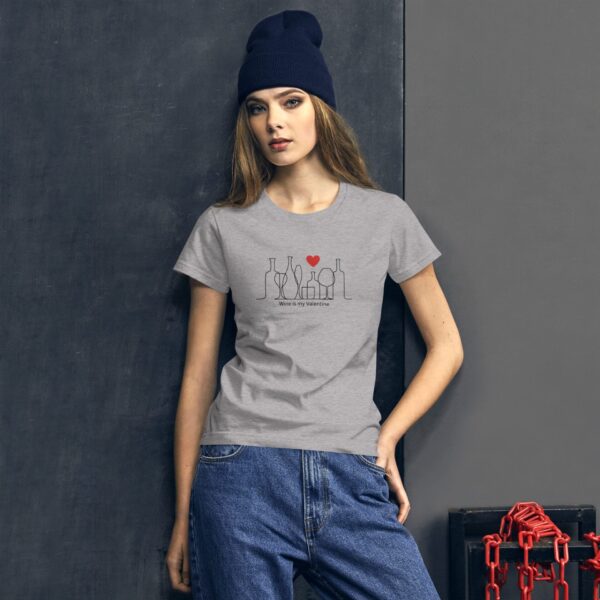A young woman stands wearing a gray t-shirt with a wine-themed graphic and the text "Wine is my Valentine." She has long hair and a navy beanie  posed against a textured wall.