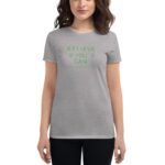 A woman stands wearing a gray t-shirt that features the green text "BELIEVE YOU CAN" and "HAPPY ST PATRICK'S DAY " with a casual and cheerful expression in a neutral background.