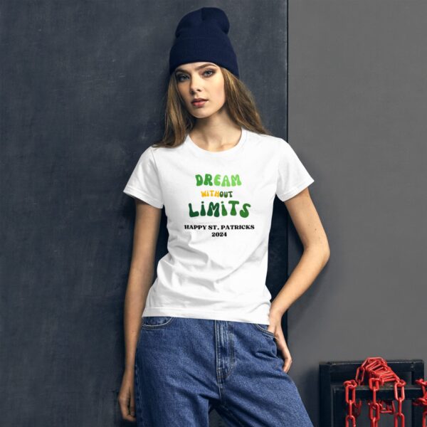 A young woman is posing while wearing a white t-shirt that reads DREAM without LIMITS HAPPY ST. PATRICKS 2024. She has long hair and a dark beanie  with a dark background and red chains nearby.