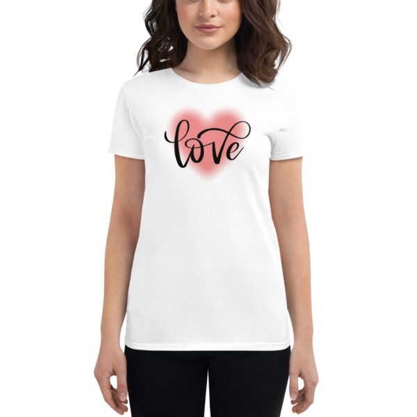 A woman is wearing a white t-shirt featuring the word love in stylish black cursive letters against a soft pink heart background  standing in a neutral setting.
