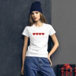 A woman stands confidently wearing a white t-shirt with red hearts spelling "LOVE" while paired with blue jeans and a dark beanie in a minimalist indoor setting with gray walls.