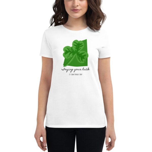 A person stands wearing a white t-shirt featuring a green four-leaf clover graphic with the text “Trying your luck It’s Saint Patrick’s Day” against a neutral background.