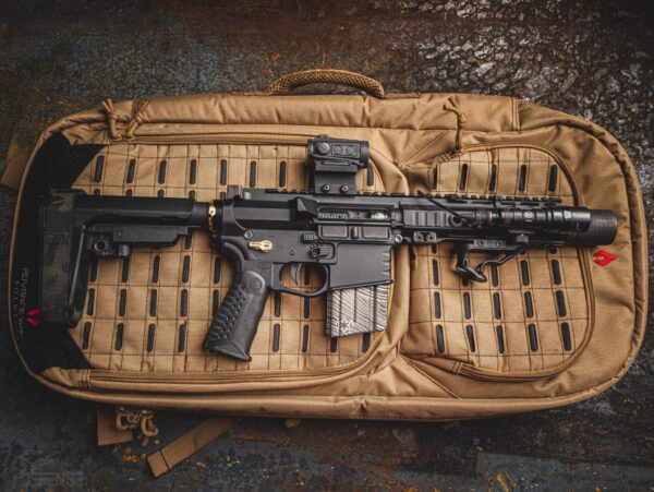 A black firearm rests horizontally atop a tan padded bag with a grid pattern The firearm features a scope and flashlight positioned on top with a textured grip and a magazine attached