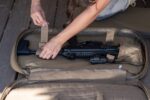 A person secures a black rifle with Velcro straps inside a large tan carrying bag on a wooden surface  ensuring the firearm is stable and protected for transport.