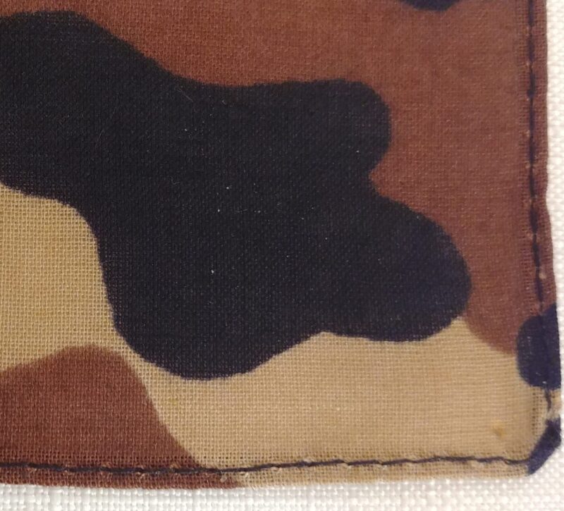 A piece of fabric featuring a camouflage pattern displays various shades of brown and black shapes intertwined against a lighter background with visible stitching along the edges.